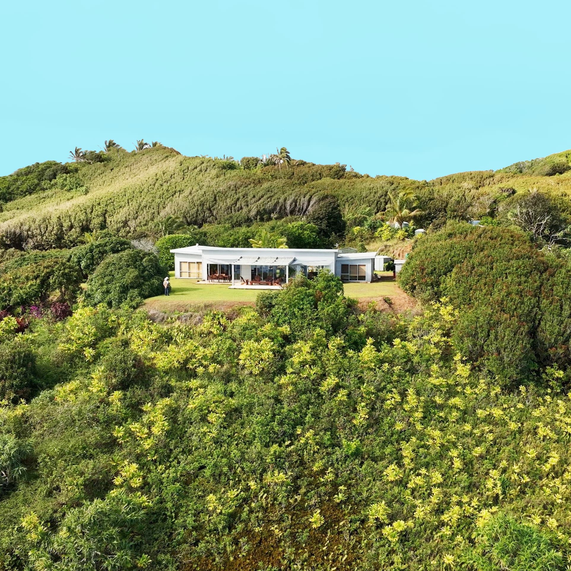 A modern white house surrounded by lush green vegetation and rolling hills under a clear blue sky