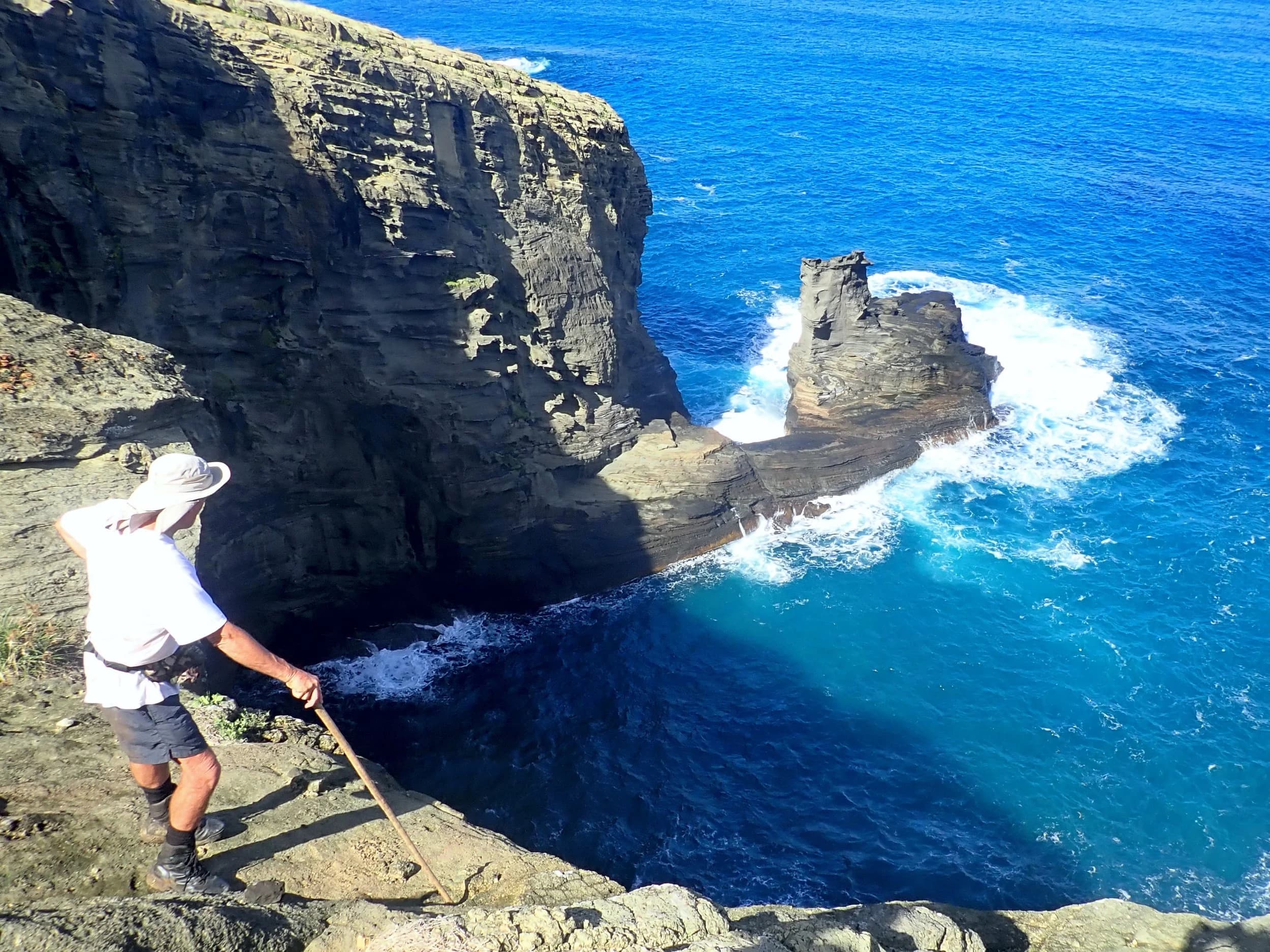 A walker in a sun hat on a rocky cliff edge above a deep blue cove