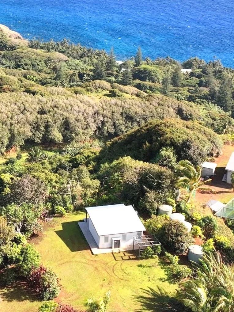 Aerial view of a small house surrounded by lush greenery near the ocean. The house has a sloping roof and nearby water tanks. The background features dense forest and the ocean with a bright blue color.